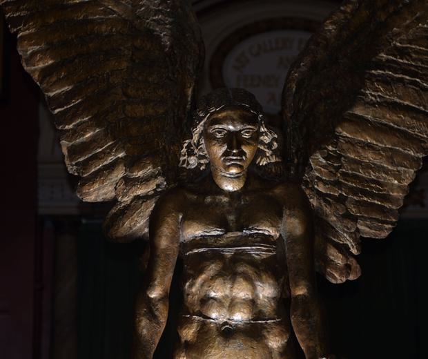 Close up of Lucifer showing his face, upper body and wings. He is partially lit and the rest of the gallery is dark.